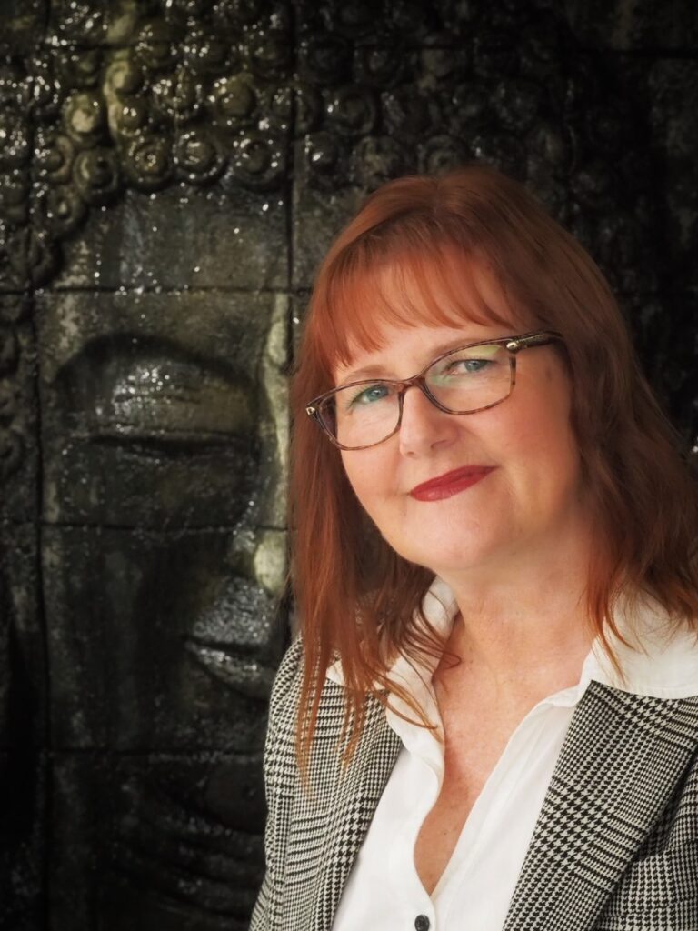 My World - Head shot of Joanne VanR. She is wearing a grey check jacket and white blouse and standing in front of a buddha ornament.
